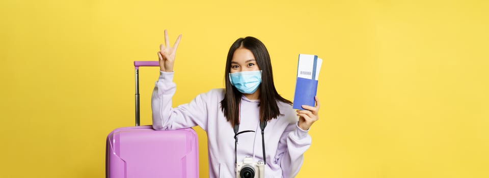 Japanese girl in medical mask, showing her passport and flight travel pass tickets, peace sign, posing with suitcase, going on vacation, yellow background photo