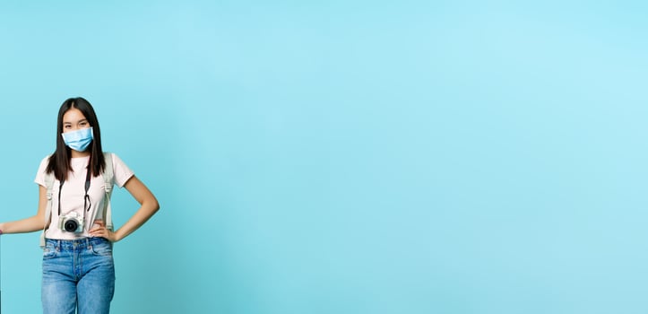 Happy asian female tourist, standing in medical face mask with suitcase and camera, going on tour, vacation abroad during covid-19 pandemic, blue background photo
