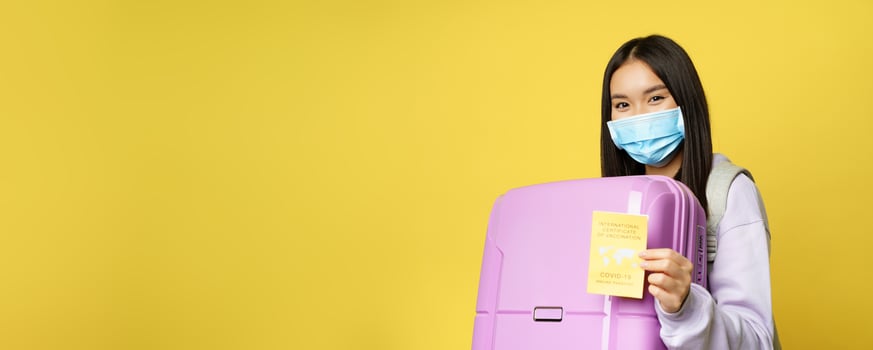 Covid-19 and tourism concept. Happy asian female tourist holding suitcase, showing international coronavirus vaccination certificate for travelling, yellow background photo