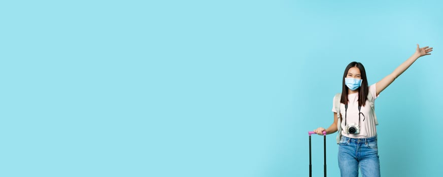 Full length of happy korean woman in face mask, standing with suitcase, raising hand up dreamy, enjoying vacation abroad, standing over blue background photo