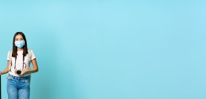 Happy asian girl traveller, tourist with suitcase and camera, going on vacation, wearing medical face mask during covid-19 restrictions, blue background photo