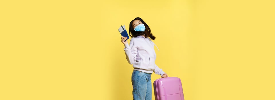 Full length shot happy asian woman jumping with suitcase and tickets on tour, holding passport, going on vacation, standing over yellow background photo