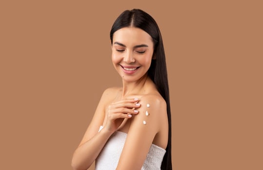 Smiling attractive shirtless long-haired brunette half-naked young woman applying body lotion or cream on shoulder, touching her smooth skin, isolated on beige studio background, copy space photo