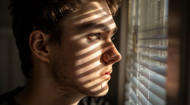 Young man looking thoughtfully through blinds with sunlight casting shadows on his face photo