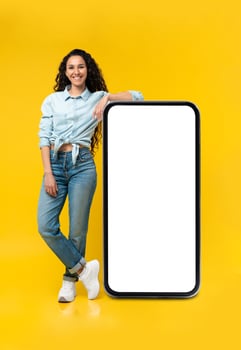 Full length of cheerful young lady standing near big smartphone with empty screen, demonstrating mockup for website, advertisement or app over yellow studio background photo