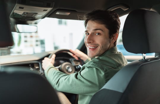 Handsome young man looking back at camera, sitting in car, parking auto, view from rear seat. Happy millennial guy buying automobile at modern dealership store, copy space photo