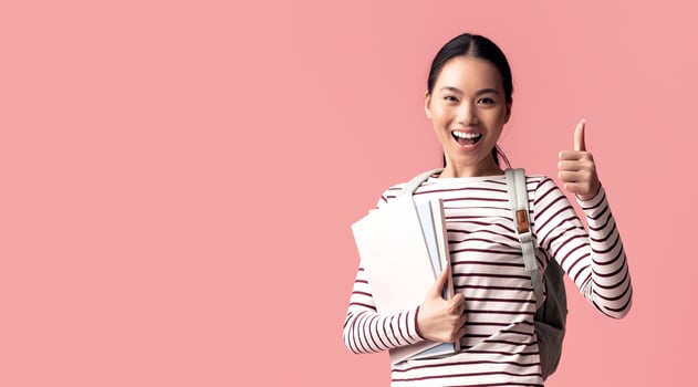 Cheerful Asian Female Student Jumping In Air With Workbooks And Showing Thumb Up, Happy Excited Korean Female Wearing Backpack And Smiling At Camera While Posing Over Pink Background, Copy Space photo