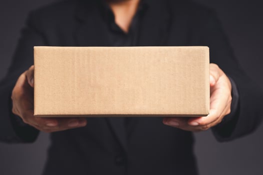 Businessman in a black suit holds and points to a cardboard box while standing on a gray background photo