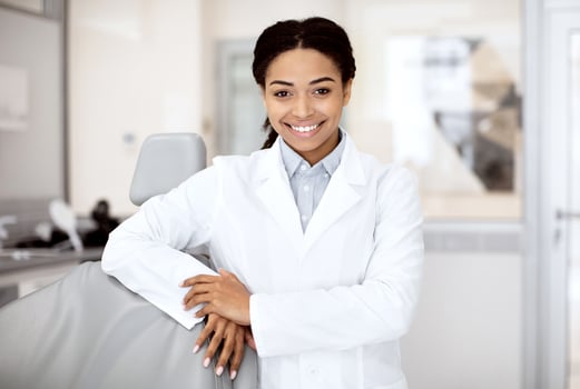 Portrait Of Young Black Female Dentist In White Coat Posing In Clinic Interior, Happy African American Stomatologist Woman Leaning On Dental Chair And Smiling At Camera, Waiting For Patient photo
