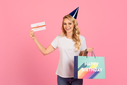 A cheerful young woman wearing a party hat holds a gift certificate and a colorful birthday bag. The backdrop is bright pink, enhancing the festive atmosphere of the celebration. photo