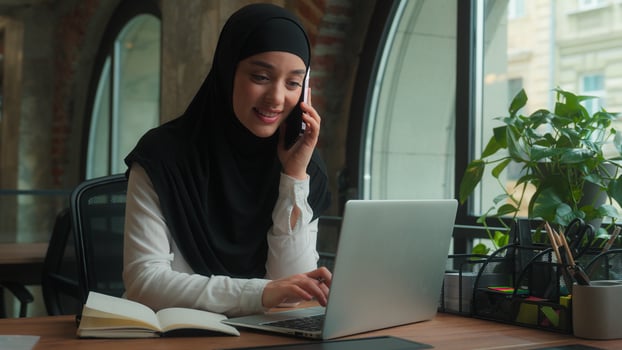 Happy muslim islamic business woman in hijab female girl Arabian multitasking businesswoman manager talk mobile phone answer smartphone call consult client online write notes typing laptop at office photo
