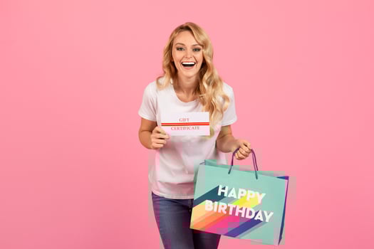 A cheerful young woman holds a birthday gift certificate while smiling broadly. She displays a colorful birthday bag, set against a bright pink background, capturing the joyful moment. photo