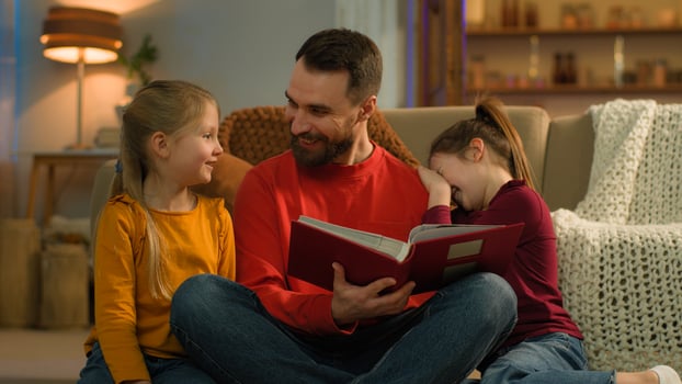 Caucasian family late night evening at home parent dad father man reading book kids watching photo album funny pictures happy cheerful amazed children little girls daughters schoolgirls enjoy learning