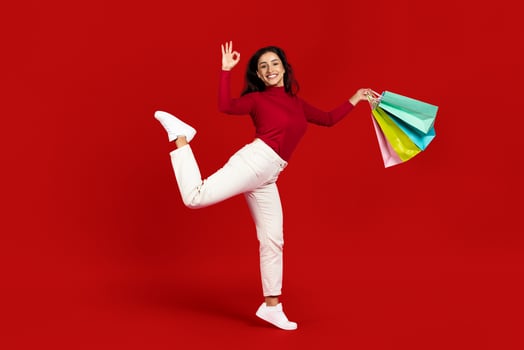 A young Caucasian woman joyfully poses against a bright red background, holding colorful shopping bags while displaying her excitement for recent purchases in a moment of celebration. photo