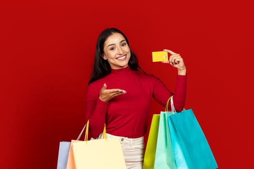 A young Caucasian woman poses cheerfully with colorful shopping bags while holding a yellow card. She enjoys the excitement of shopping and consumerism in a striking red setting. photo