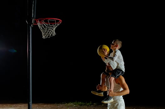 A father joyfully lifts his son for a basketball shot under the night sky at a local court photo