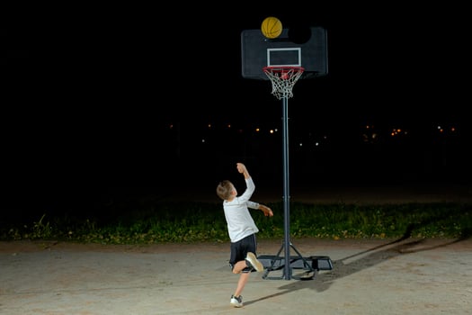 A child shoots a basketball at a hoop during nighttime practice in an outdoor playground with city lights in the background photo