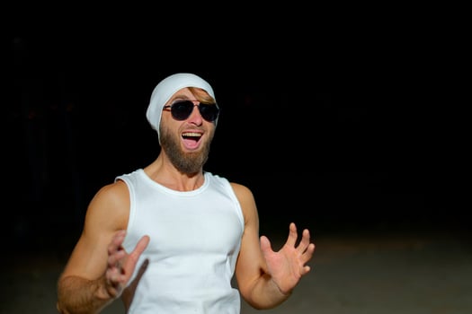 A man wearing a tank top, sunglasses, and a beanie poses confidently at night against a dark background photo