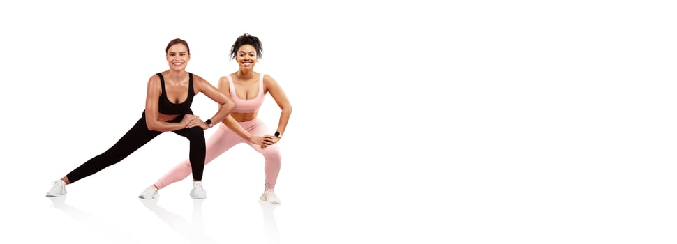 Two women perform side lunges together in a bright, minimalistic indoor space. They wear matching athletic attire and demonstrate camaraderie while focusing on their fitness routine. photo