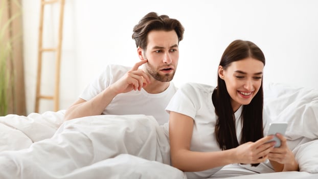 Jealousy. Suspicious Husband Reading Wife's Messages Behind Her Back While She Texting In Social Media In Bedroom At Home. Man Checking Cheating Girlfriend's Phone. Marriage Problem, Infidelity photo