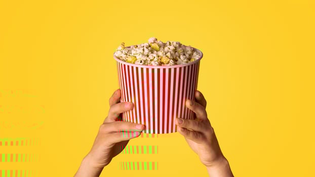 Unrecognizable man holding striped paper bucket with popcorn on orange background, closeup. Panorama photo