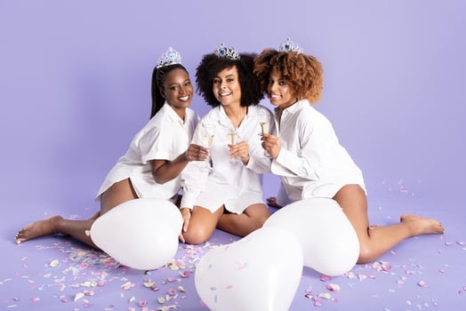 Three young African American women sit together on a purple background, wearing crowns and sharing laughter while holding drinks. photo