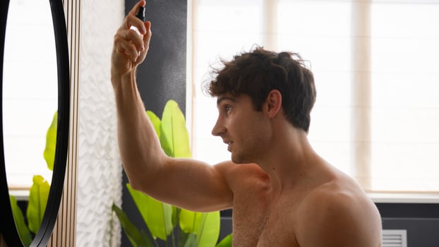 Caucasian naked man guy male in bathroom morning beauty routine procedure looking at mirror reflection apply spray hair spraying thermal water hairstyle care alopecia baldness prevention after shower photo