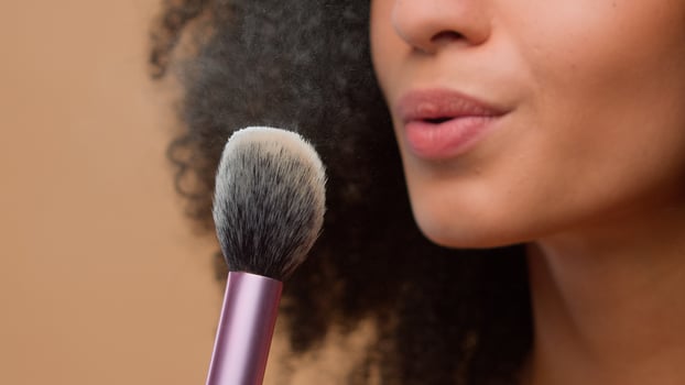 Cosmetic make-up beauty concept close-up cropped view unrecognizable African American woman unknown model girl holding brush blow away dust apply skin tone powder at beige studio background skincare photo