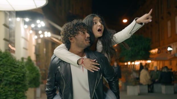 Indian Arabian couple love guy holding girl on back piggyback woman man boyfriend girlfriend walking night evening city looking at beautiful urban architecture pointing excited tourist travel dating photo
