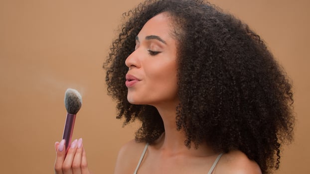 Close up portrait African American ethnic woman model girl holding brush blow away dust apply skin tone powder to nose smiling at camera at beige studio background beautiful lady cosmetics make-up photo