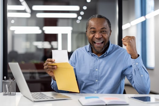 Mature African American businessman celebrates success at office desk with laptop and open envelope, displaying joy and accomplishment. Image conveys excitement, achievement, and career progress. photo