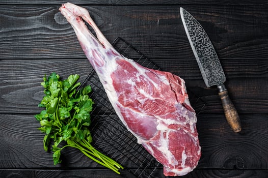 Fresh Raw whole lamb mutton leg thigh on kitchen table with herbs. Black Wooden background. Top view photo