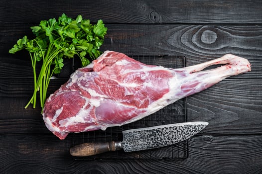 Fresh Raw whole lamb mutton leg thigh on kitchen table with herbs. Black Wooden background. Top view photo