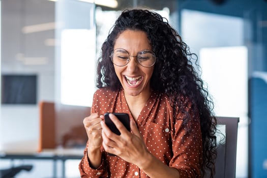Young beautiful hispanic woman with curly hair inside office celebrating victory success, businesswoman received win notification on phone, using smartphone app at workplace. photo