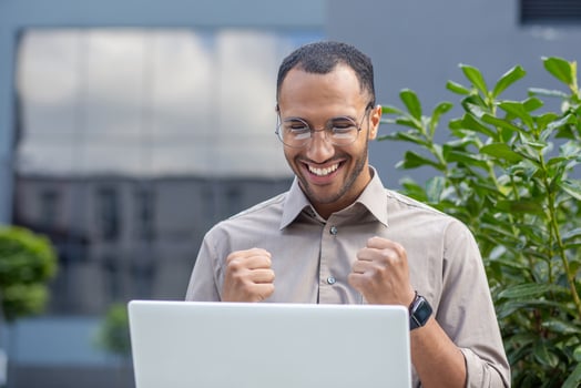 Close-up photo of a happy young hispanic man sitting outside an office center on a bench and smilingly looking at a laptop screen showing a victory gesture with his hands.