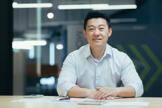Portrait of a young Asian freelancer, designer, architect. He sits at the desk in the office, holds the phone, works with documents, looks into the camera, smilling. photo