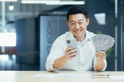 A happy young Asian man sits in the office at the table, holds a phone and cash money in his hand. A smiling man dials the phone, rejoices at a monetary win, lottery photo