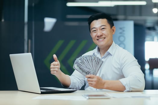 Portrait of a young handsome Asian businessman, boss, SEO millionaire. He sits in the office at the table with a laptop, holds money in cash in his hands, smiles at the camera, points a super finger. photo