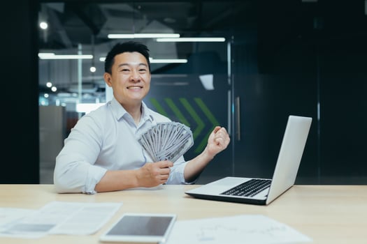 Portrait of a happy Asian man working in the office on a laptop, holding a fan of cash, dollar bills in his hand. shows a victory gesture with his hand to the camera, celebrates. photo