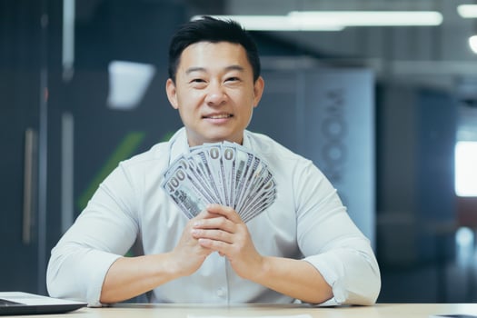 A happy young Asian businessman, freelancer, manager, office worker holds a cash of money in his hands and waves it. He is happy, points to the camera. Sitting at a desk in a modern office photo