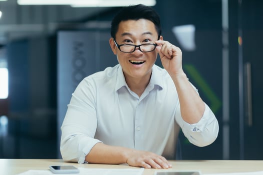 Portrait of a happy young Asian man sitting in the office at the table with phone and documents, holding glasses in his hand, looking down, smiling, looking at the camera. photo