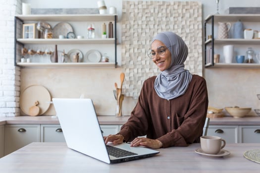 Young beautiful Muslim woman in hijab working remotely from home, using laptop while sitting in kitchen at table, woman in glasses smiling and happy with work. photo