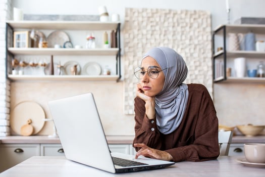 Bored young muslim woman wearing hijab sitting in the kitchen at home, resting her head on her hands, looking sad and moody at laptop. photo