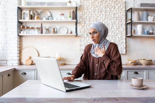 A young Arab woman in hijab is working, studying at home using a laptop. Holds her hands to her chest, looks at the monitor in shock, received bad news, feels a panic attack, worries, severe stress. photo