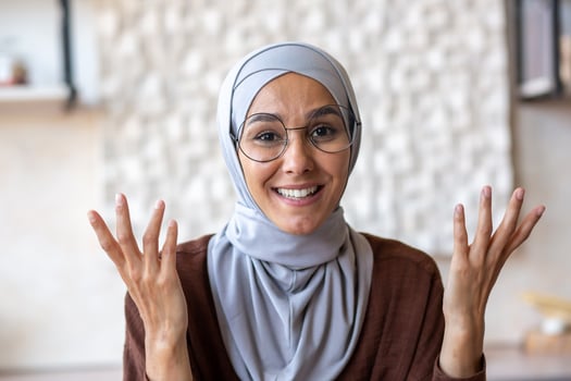Close-up photo. Arab young woman in hijab and glasses talking to camera from home. Gesturing with hands, smiling. photo