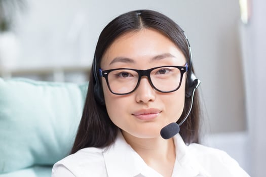 Close-up portrait photo of Asian female call center worker and tech support worker, looking at camera and smiling, using headset for call.