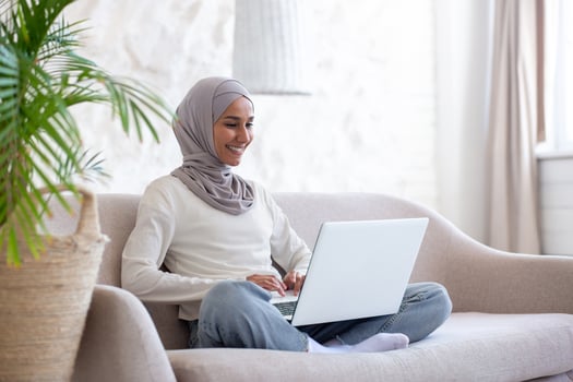 Smiling young Muslim woman in hijab sitting on couch at home in lotus yoga position and working remotely online on laptop. photo