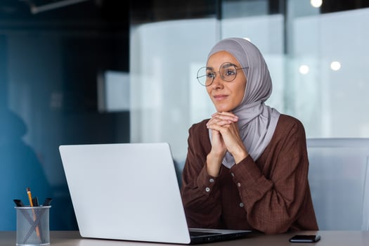 Portrait of a young Arab woman in a hijab and glasses sitting in the office at a table, working on a laptop. She rested her head on her hands. Thoughtfully and dreamily looks ahead, out the window. photo