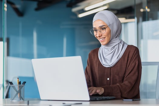 Happy and successful business woman in hijab working with laptop inside modern office building, muslim woman in glasses smiling and happy with work achievement. photo