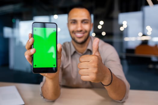 Happy man sitting in office holding phone with green screen and showing thumb up, phone in foreground. photo